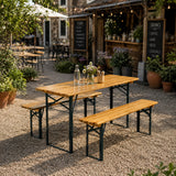 A rectangular outdoor picnic table with a light wood top and black metal frame, accompanied by two matching wooden benches, set on a gravel patio in front of a rustic wooden bar.